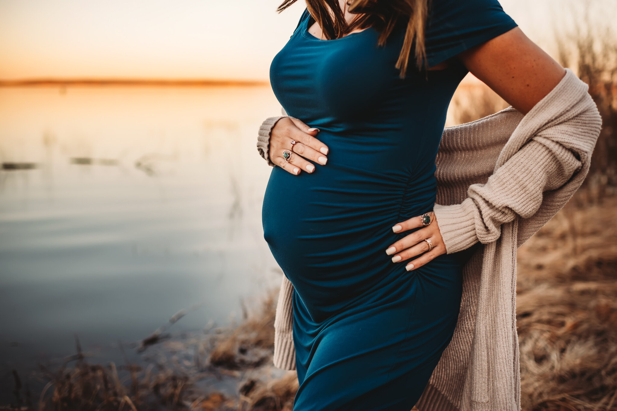 central Illinois family photographer. Mom stands on riverbank with sunset, holding pregnant stomach
