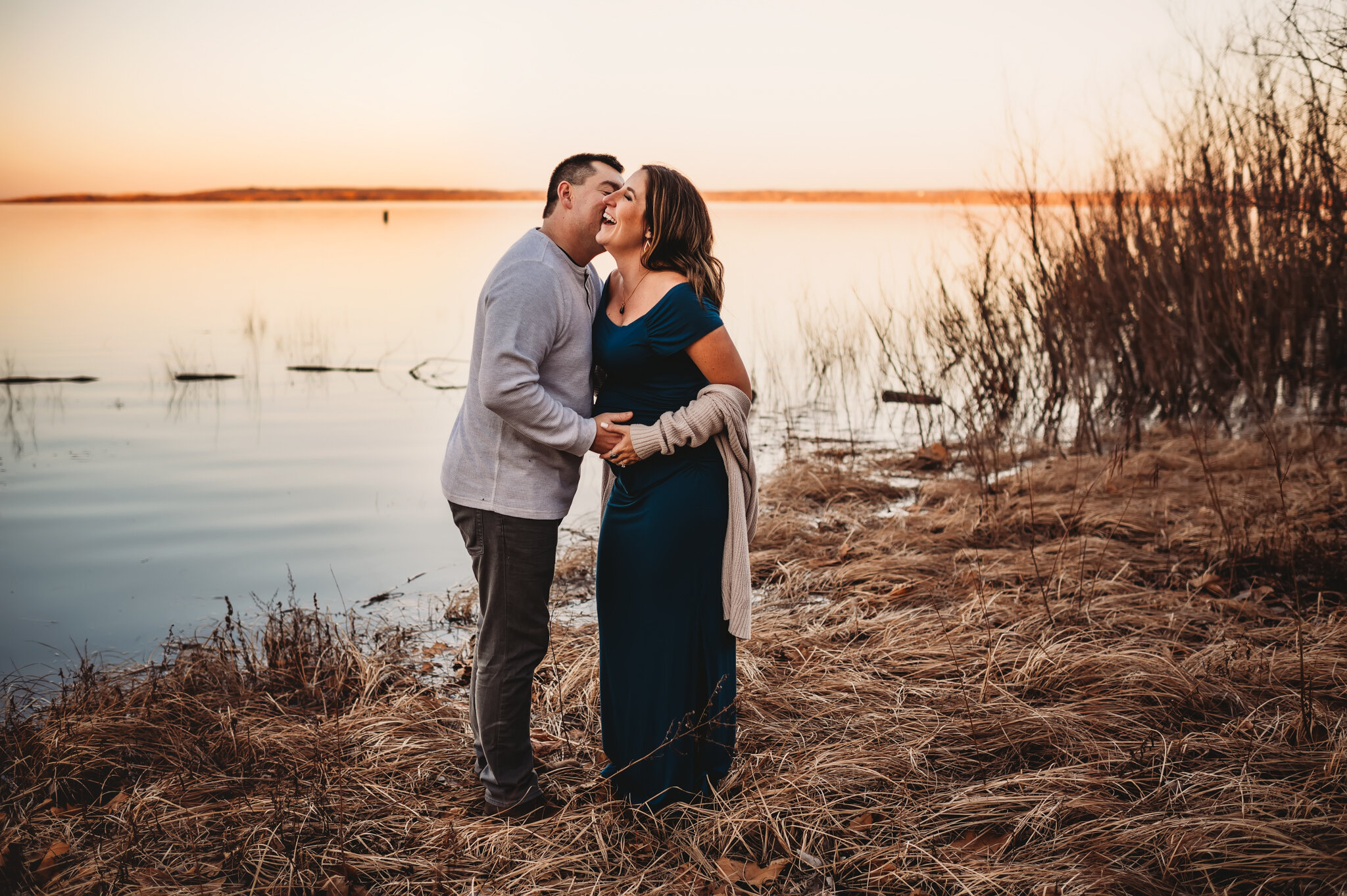 Peoria Illinois maternity photos, couple stands near river kissing and laughing during maternity photoshoot