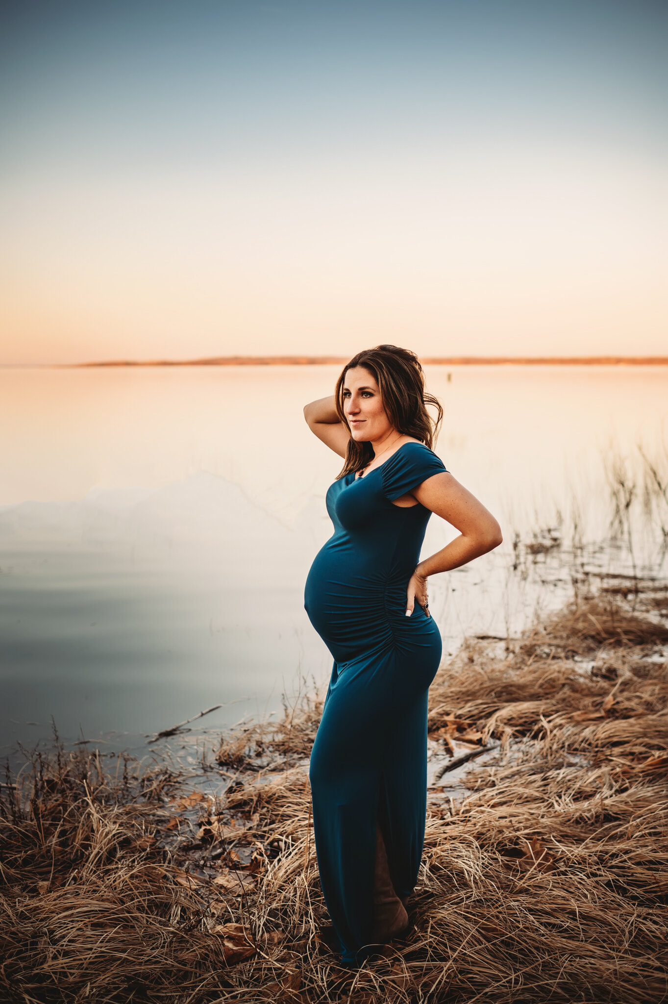 peoria IL maternity, glamorous pregnant mom poses near water with sunset wearing blue maternity dress
