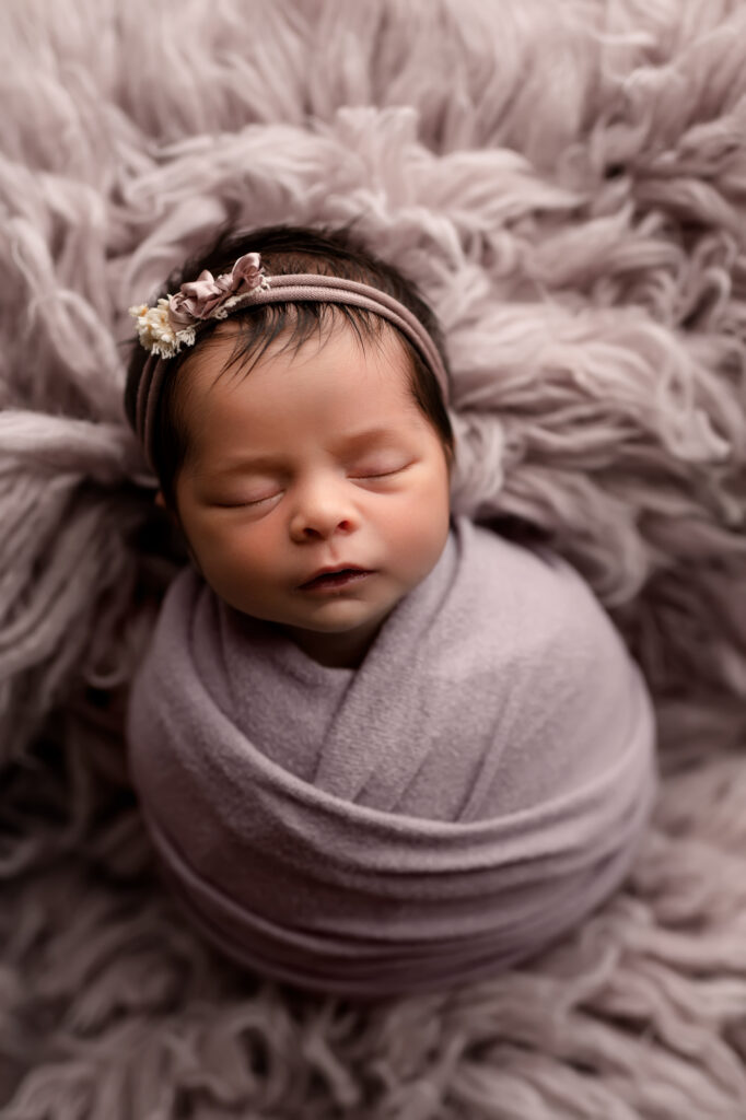 Newborn baby girl lays on pink fur in pink and purple wrap and bow