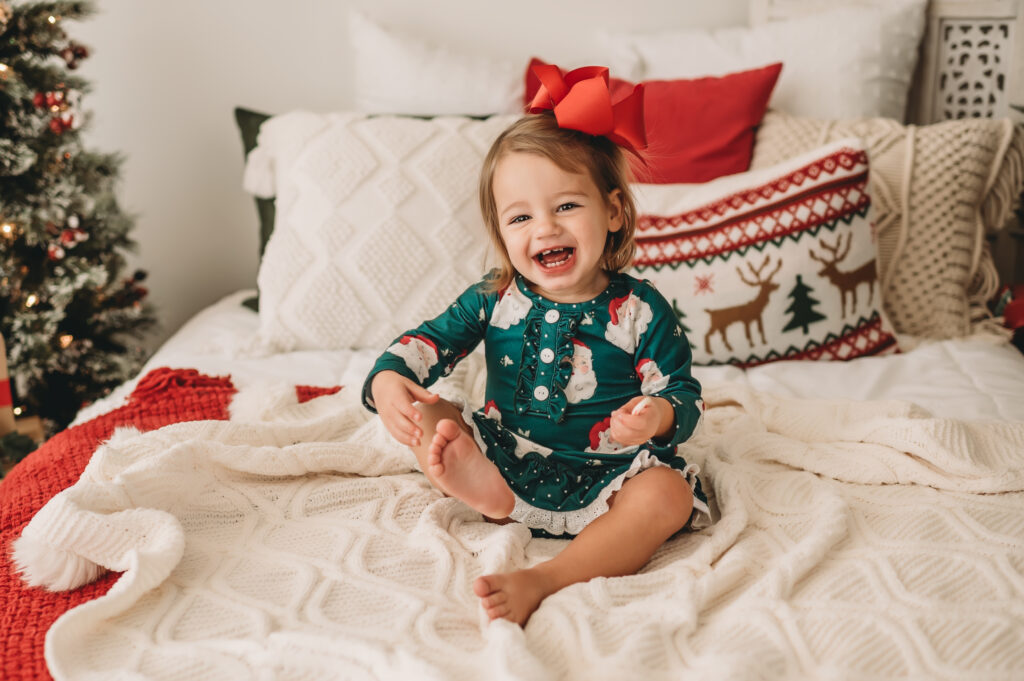 Little girl sits on bed and giggles wearing Santa jammies and red bow