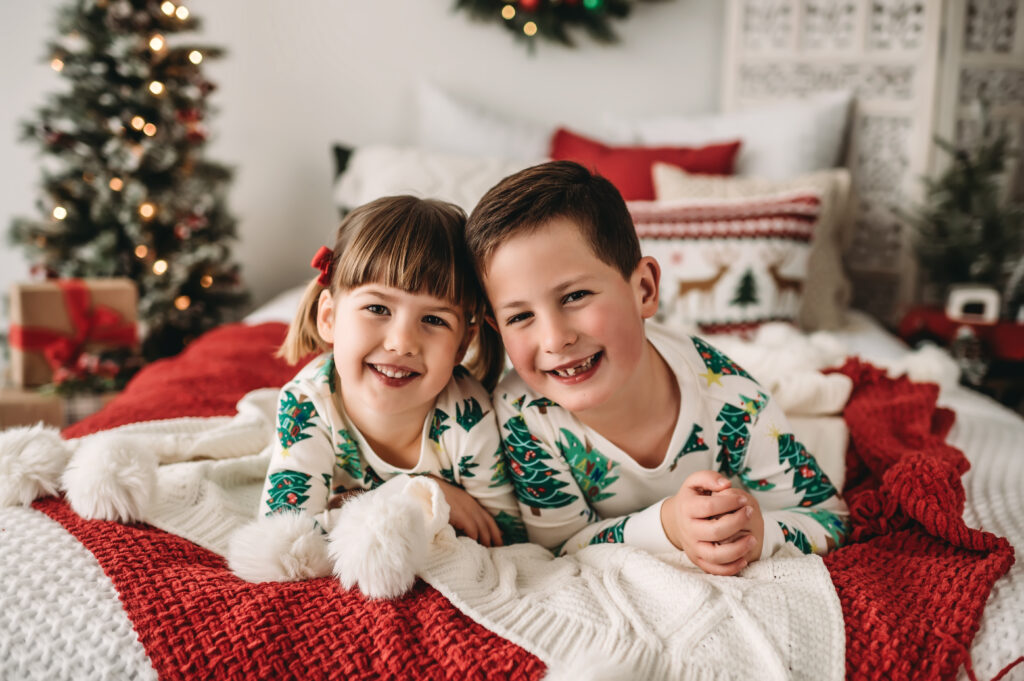 siblings sit on bed together in Christmas tree jammies