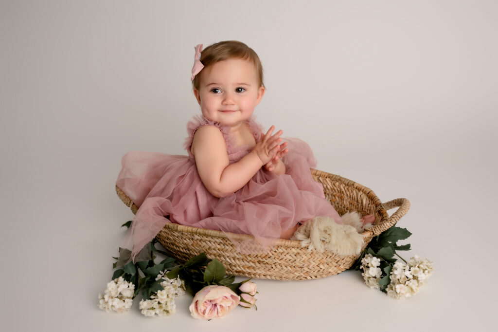 baby girl in pink dress sits in basket with flowers