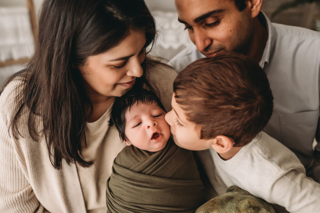 family poses together. Older brother kisses newborn while he yawns