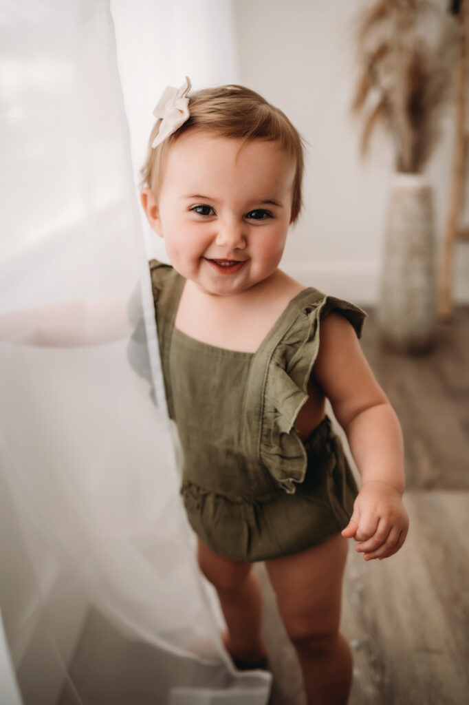 baby girl in olive green outfit stands near curtain